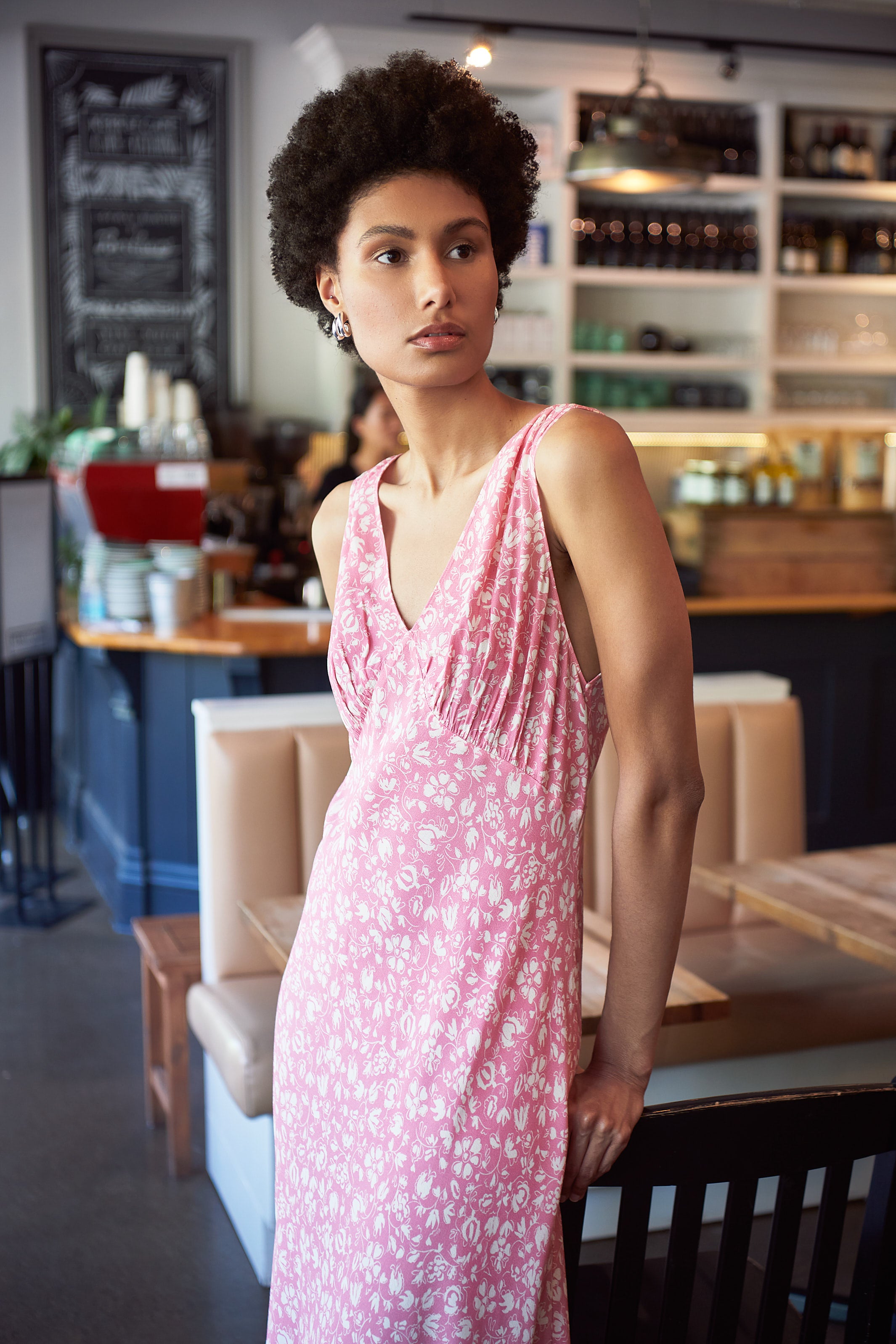 A woman wearing the Sandrine Sleeveless Pink Midi Dress by RIXO leans against a table in a cafe